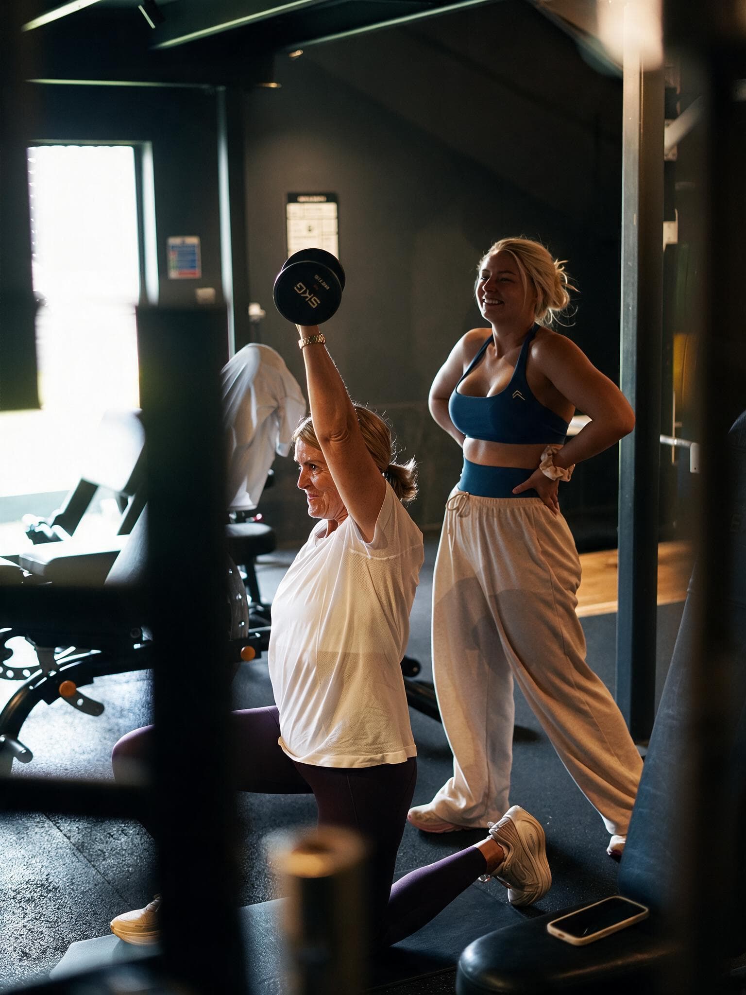 A client presses a dumbbell overhead mid-lunge while Beth stands behind her